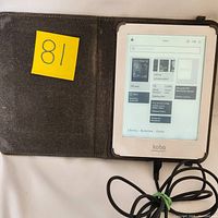 Kobo EReader inside brown protective case, displaying eBook library with a few Stephen King titles loaded, charging cable connected.