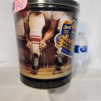 Front and side view of the unopened cylindrical Cheetos can featuring Ken Danby's 'Lacing Up' artwork, with a water bottle for scale.