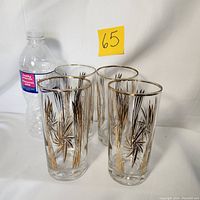 Four vintage glass tumblers approximately 5.5 inches tall next to a water bottle for scale. Glasses have gold gilt starburst design and gold rims, with some wear.