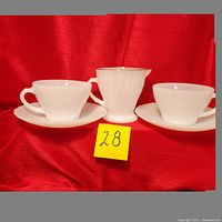 Two white swirl patterned milk glass teacups with matching saucers flanking a gold-rimmed milk glass creamer, all placed on red fabric.