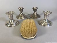 Overview of 2 pairs of weighted silver candleholders and a silver handled brush with bristles, arranged on neutral surface