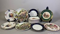 Photo showing an assortment of decorative plates and dishes including fruit-themed, cottage landscape, cobalt blue rimmed, and floral designs.