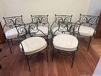 Six wrought iron chairs with white cushions arranged in a row on hardwood floor against plain wall