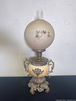 Full view of lamp showing frosted floral globe atop the ornate base with brass cherub accents and glass chimney.