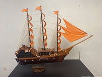 Full view of the model ship showing three masts with orange fabric sails and rigged string details on the hull and deck, resting on a wooden stand.