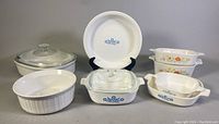 Seven vintage CorningWare casserole dishes with some glass lids. Blue Cornflower and orange floral patterns visible. Various sizes and shapes including round and rectangular.