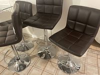 Four brown faux leather vinyl bar stools with chrome bases arranged in a room, showing their seats and bases.