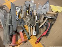 Wide view of assorted concrete tools including multiple trowels, scrapers, and wire brushes with various wooden and plastic handles.