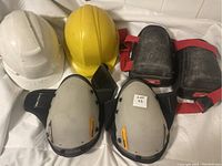 White and yellow hard hats placed alongside two pairs of knee protectors, one with black and red straps and the other with black foam padding.