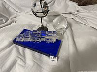 Three crystal items on white fabric including a suspended NTBA sphere with metal stand, a truck shaped crystal on a blue rectangular base, and a clear crystal sphere beside them.