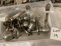 Plastic bin with multiple metal door knobs and lever handles loosely placed inside. Various parts and locking mechanisms visible.