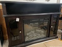 Straight-on photo of electric fireplace in dark wood mantel with closed glass door cabinets on each side, showing artificial logs inside fireplace insert.