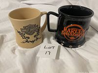 Two Harley Davidson officially licensed mugs, one beige with black tribal logo, one black with orange logo and silver studs.