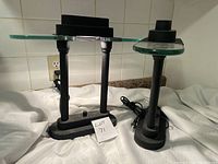 Two electric devices on a table, one being a lamp missing bottom bulb glass cover and the other a heater with metal vented top.