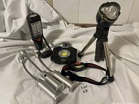 Five portable lamps consisting of three silver magnetic base lights, one handheld black and yellow lamp, one black tripod lamp, and one headlamp lying on white cloth.
