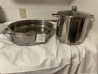Front view of a large stainless steel stock pot with glass lid and a stainless steel roaster pan with lid resting on a white cloth background.