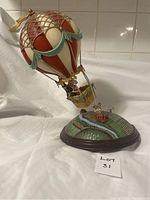 Side view of musical hot air balloon figurine showing painted metal balloon and detailed base with building and river.