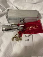 Oxygenics shower head lying in front of silver metal case and red velvet bag on white fabric background with Lot 62 note visible.