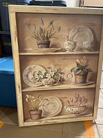 Photo of framed print titled 'Pritemps 1' by Fabrice de Villeneuve with depiction of potted plants and decorative plates on shelves.