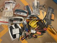 Overview photo of lot showing various tiling tools including notched trowels, scrapers, pliers, suction handle, grout spacers, rubber mallet and hand tools scattered on surface.