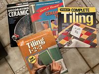 Four ceramic and tiling instructional books arranged on a surface, showing covers clearly: Black & Decker's Decorating Ceramic, Creative Homeowner Press's Quick Guide Ceramic Tile, Stanley's Complete Tiling, and The Home Depot's Tiling 1-2-3 2nd Edition.