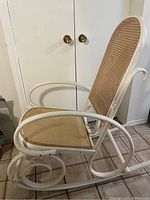 Side angled view of the white wooden rocking chair frame with woven wicker backrest and seat base showing the curved armrests and rocker supports.