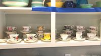 Twenty sets of assorted china teacups and saucers displayed on shelving, showing various floral and decorative patterns with gold trim and English styles.