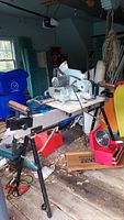 Side view of the Delta compound miter saw on the metal stand with a blue tarp and surrounding garage clutter.