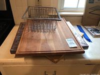 Photo showing three wooden cutting boards stacked, one new in packaging, with metal wire dish strainer on top.