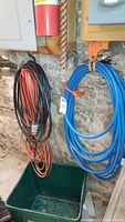 Three coiled utility extension cords and one blue Amflo air compressor hose hanging against a stone wall in a basement.