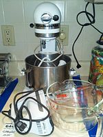 White KitchenAid heavy duty lift stand mixer with attached stainless steel bowl, visible electrical cord, whisk, beater, and dough hook inside bowl, and two clear Pyrex measuring bowls in front.