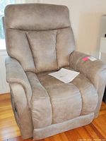 Front-view of the light brown synthetic suede upholstered recliner with padded cushions and seams visible along armrests and backrest. A controller and paper manual rest on the seat.