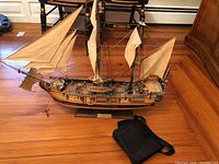 Wooden detailed model ship with sails and rigging on display on wooden floor, black binocular case on the floor next to it