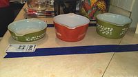 Photo showing full set of three Pyrex Cinderella bowls: two olive green Spring Blossom patterned bowls and one orange Autumn Harvest Wheat bowl with chip visible on handle.