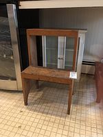 Photo of a wooden shelf unit and smaller wooden table with visible wear, next to a white shelving unit with frosted glass doors