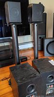 Two Samsung cube surround speakers mounted on tall matte-black stands alongside a subwoofer and TV stand in background.