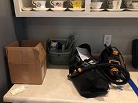 Overview of lot items including cardboard box, green cleaning caddy with cleaning products, McCulloch steam cleaner with hose and handle, black folded air conditioner cover, and fold-up broom.