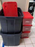 Two large black plastic storage bins, one with red lid removed and placed inside top bin, next to red storage drawer unit with three transparent drawers with silver handles