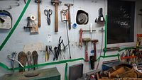 Wide view of assorted hand tools hanging on a wall including hammers, mallets, multiple pliers, scissors, tape, and other small tools near a green toolbox.
