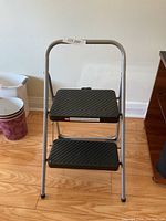 Full view of 2-step ladder showing silver metal frame and two wide black textured steps with rubber foot grips.