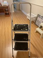 Front view of the 3 step ladder showing white metal frame and black steps with paint splatter