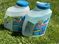 Two large plastic containers of Alaskan Degelcant Premium ice melter on grass, showing front labels and blue lids.