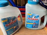 Two 5 kg opaque white plastic containers of Alaskan Premium Ice Melter with blue lids and handles in a storage setting.