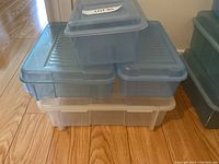 Several assorted plastic storage bins stacked on a wooden floor, mostly translucent blue with lids, including one large translucent white bin at the bottom.