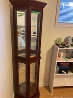 Curio cabinet showing front and side glass panels, wooden frame in reddish-brown finish, multiple glass shelves inside, set against a beige wall with window nearby.