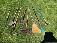 Full view of 8 garden tools laid on grass including fork, shovels, rakes, brooms and cultivator.