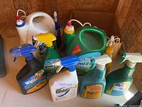 Photo showing various weed and pest control spray bottles and large liquid containers clustered together on a wooden surface.