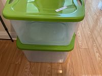 Two large translucent plastic storage bins stacked vertically on hardwood flooring, showing green snap-on lids with built-in handles. Bins are clear so contents would be visible.