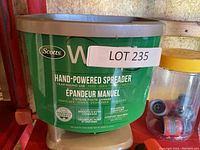 Green Scotts branded hand-powered spreader with brown top, labeled for year-round use including fertilizer and seed spreading, alongside a jar of hose connectors.