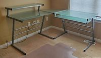 View of the three-piece glass and metal office desk set arranged in an L-shape in a corner of the room, showing main desk, corner piece, and side desk with shelf.